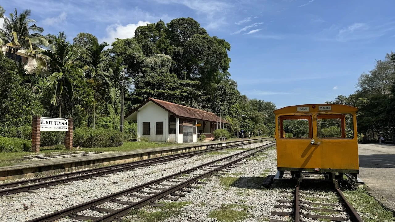 Schienen, links ein helles Gebäude im Hintergrund , rechts vorne eine Art kleine Waggon