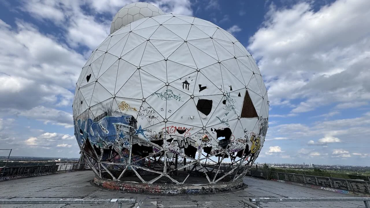 Große weiße Kuppel, mit Graffiti, teilweise ist die Hülle kaputt, im Hintergrund Blick auf Berlin